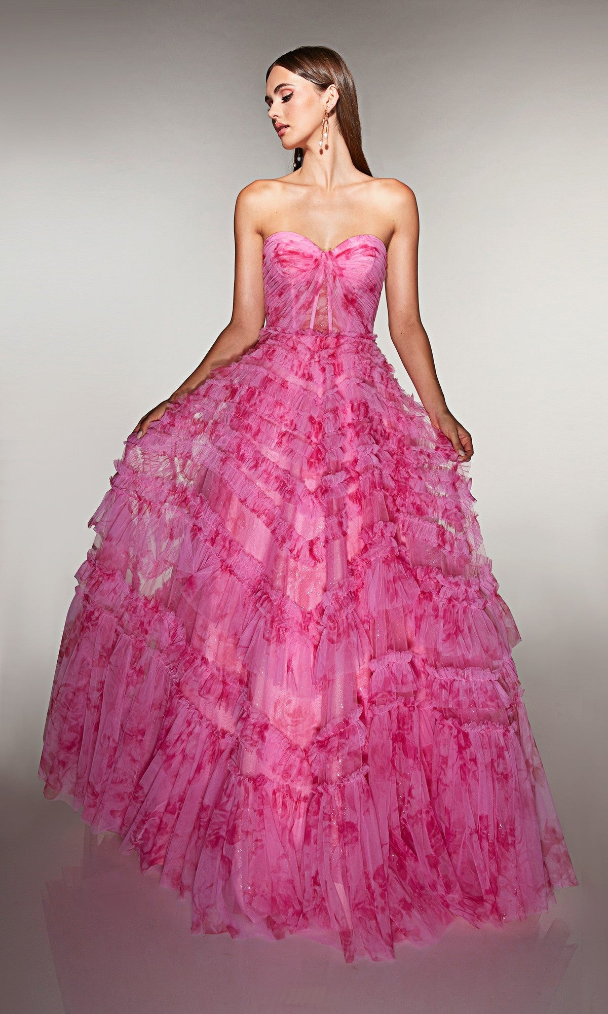 A woman stands against a neutral background wearing the Alyce 62164 long prom dress in bubblegum—a strapless gown with a corset bodice, floor-length tiered ruffle skirt, and floral patterns.